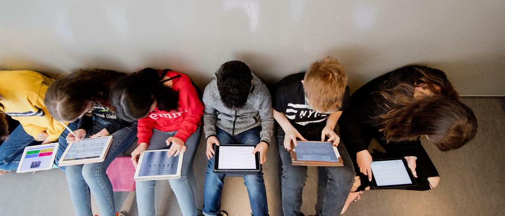 Schüler und Schülerinnen sitzen an einer Wand auf dem Fußboden und arbeiten mit Tablet-Computern.