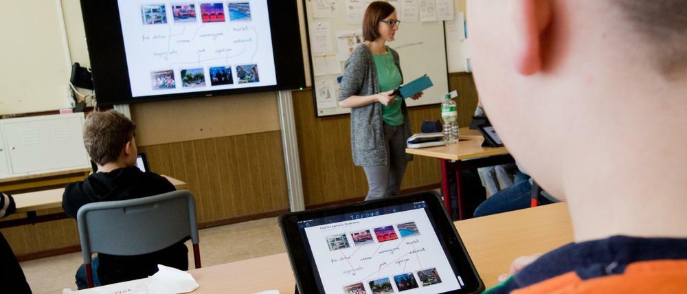 Ein Schüber sitzt vor einem Programmmenü auf seinem IPad in der Klasse, vorne an der digitalen Tafel erscheint dasselbe Bild.