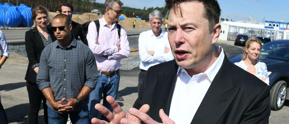 Elon Musk steht gestikulierend am Rande einer Baustelle, um ihn herum stehen Männer und Frauen, die ihm zuhören.