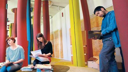 In einem künstlerisch gestalteten Raum sitzen oder stehen drei junge Menschen und lesen in Bilderbüchern.