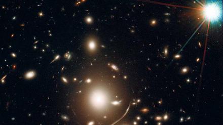 Kosmische Lichtspiele. Der Galaxienhaufen „Abell 383“ wirkt wie eine Linse, die das Licht weit entfernter Galaxien bündelt und verstärkt. Die Bilder sind jedoch verzerrt. Das zeigt sich besonders an den leuchtenden Bögen um die elliptische Galaxie (unten links). 