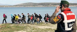 Eine Gruppe von Menschen mit Gepäck geht an einer Meeresküste entlang, im Vordergrund steht ein Polizist.