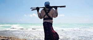 Ein Mann, der ein Gewehr über den Schultern trägt, steht auf einem felsigen Ufer und schaut über das Meer.
