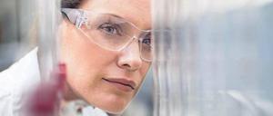 Eine junge Frau mit Schutzbrille ist hinter gläsernen Laborgeräten zu sehen.