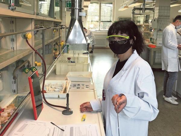 Eine junge Frau mit schwarzem Mundschutz und gelber Schutzbrille steht in einem Chemie-Labor am Bunsenbrenner.