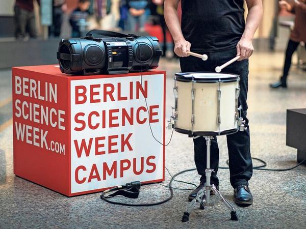 Neben einem Kubus mit der Aufschrift Berlin Science Week Campus steht ein Trommler mit Instrument.