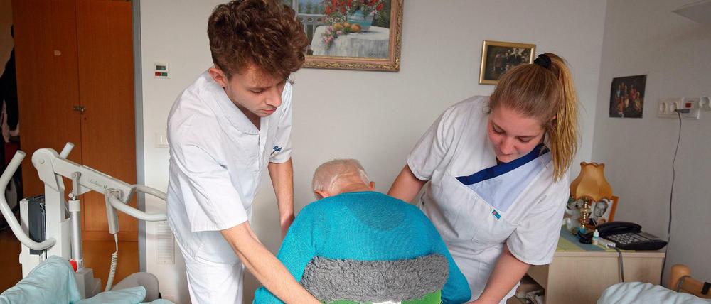 Zwei Auszubildenden in der Altenpflege helfen einem hochbetagten Bewohner beim Aufstehen.