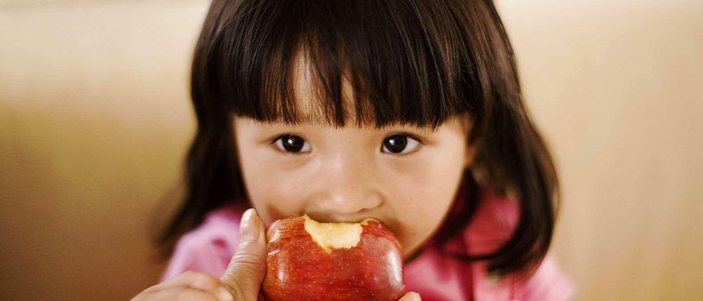 Gesunder Genuss. Ein kleines Mädchen beißt in einen Apfel, den ihr die Hand einer Erwachsenen hinhält.