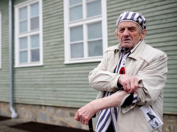 Ein KZ-Überlebender steht vor einer rekonstruierten Baracke und zeigt seine in den Arm tätowierte Häftlingsnummer.