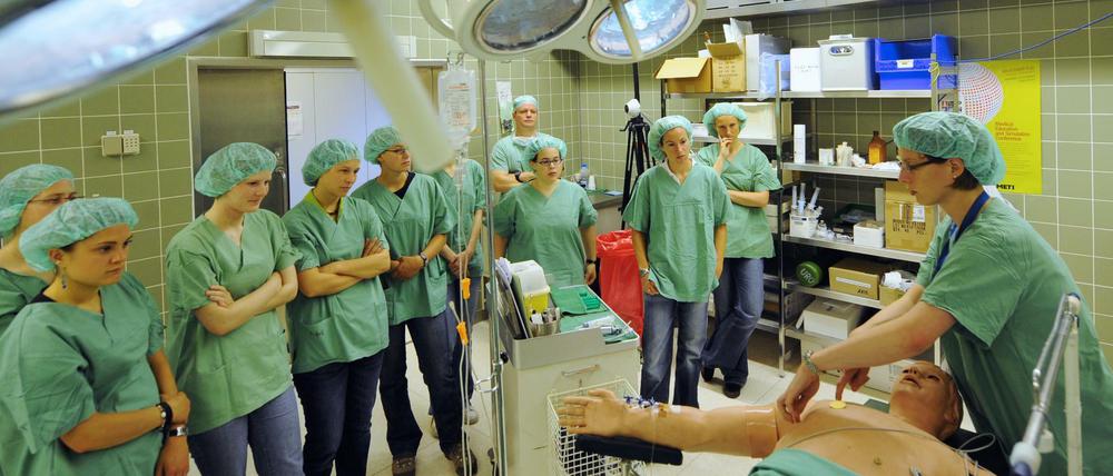 Studierenden in grünen Kitteln stehen in einem Operationssaal um eine Übungspuppe herum.