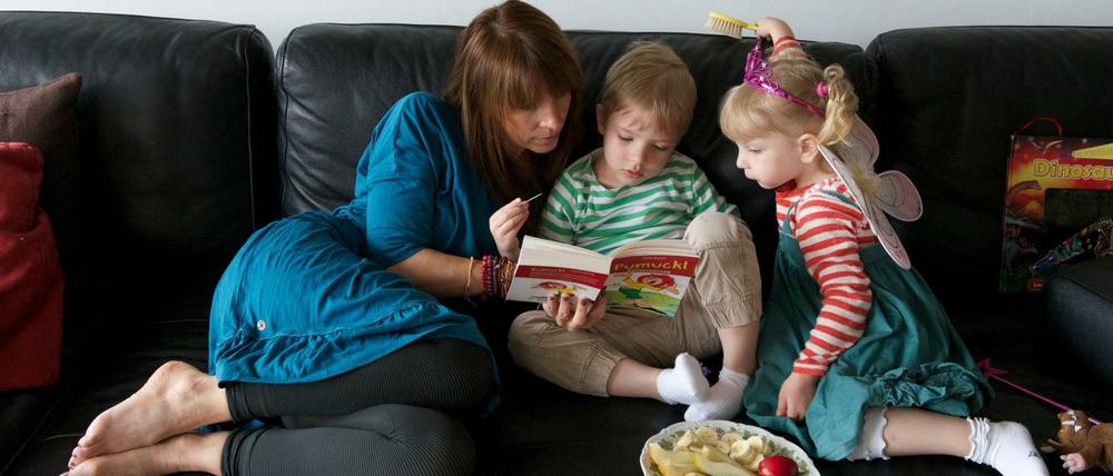 Eine Frau sitzt mit zwei Kindern auf einem Sofa und schaut mit ihnen in ein Buch; ein Obstteller steht bereit.