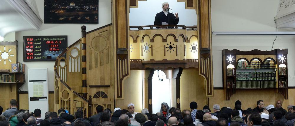 Ein Imam steht auf einer Empore und predigt, unten im Bild betende Männer.
