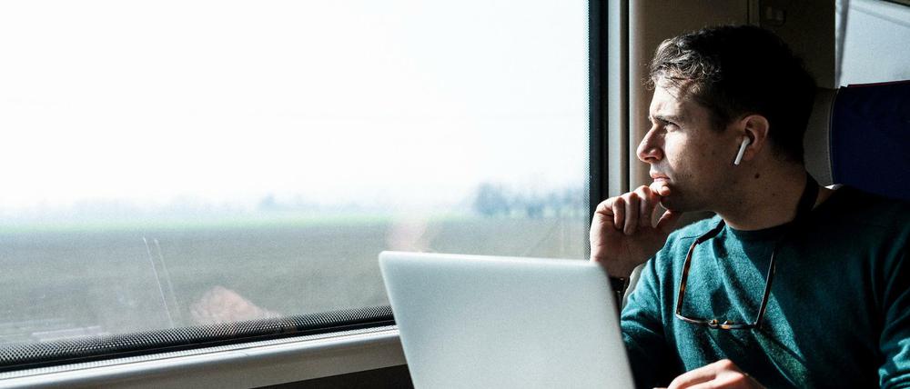 Ein junger Mann sitzt im Zug an einem Laptop und schaut nachdenklich aus dem Fenster.