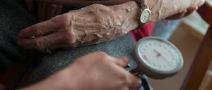 Eine Pflegekraft misst bei deiner Bewohnerin eines Seniorenheims den Blutdruck. Von beiden sind Hände und Unterarme zu sehen.