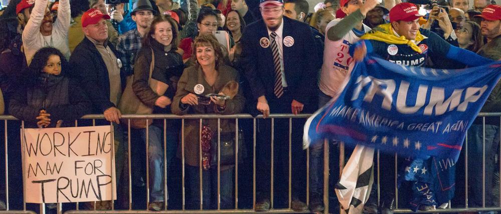 Menschen, die Transparente mit der Aufschrift Trump hochhalten, stehen jubelnd hinter einer Absperrung. Im Vordergrund ist ein Schild mit der Aufschrift "Working Man For Trump" zu sehen.