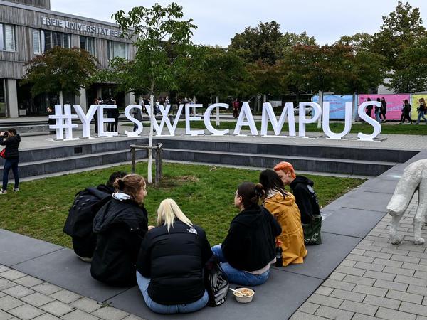 Studierende sitzen auf dem FU-Campus auf einer Mauer, im Hintergrund eine Skulptur aus dem Schriftzug Yes we Campus.