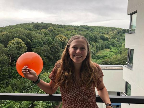 Eine junge Frau steht lachend auf einem Balkon und hält einen orangen Luftballon in der Hand.