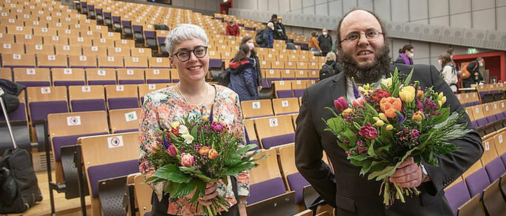 Eine Frau und ein Mann stehen mit Blumensträußen in der Hand in einem Hörsaal.