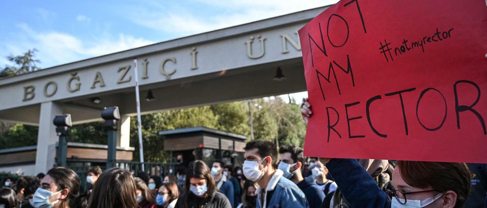 Studierende mit Mundschutz stehen vor einem Universitätsgelände, einer hält ein Plakat mit der Aufschrift Not my rector hoch.