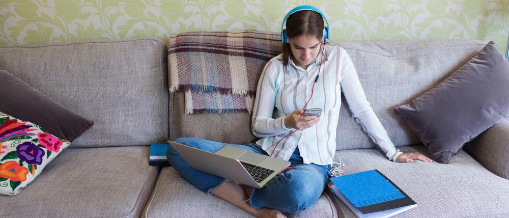 Eine junge Frau sitzt auf einem Sofa, hat ihr Laptop auf dem Schoß, ein Smartphone in der Hand und trägt Kopfhörer.