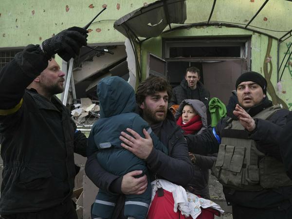 Angriff auf Zivilbevölkerung. Ein Mann trägt sein Kind aus einer durch einen Angriff beschädigten Geburtsklinik in Mariupol.